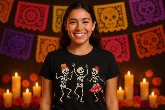 Mujer joven sonriendo mientras usa una playera negra personalizada con diseño de calaveras bailando, fondo decorado con velas y papel picado típico del Día de Muertos.